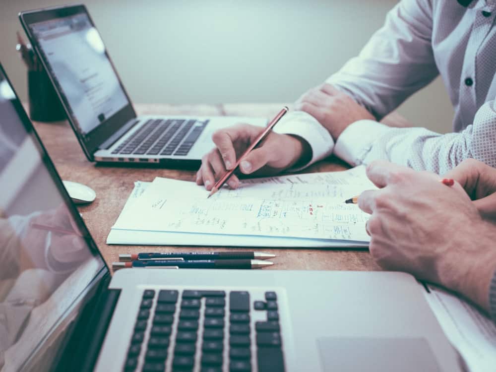 Dos personas revisando documentos con portátiles y cuadernos sobre la mesa